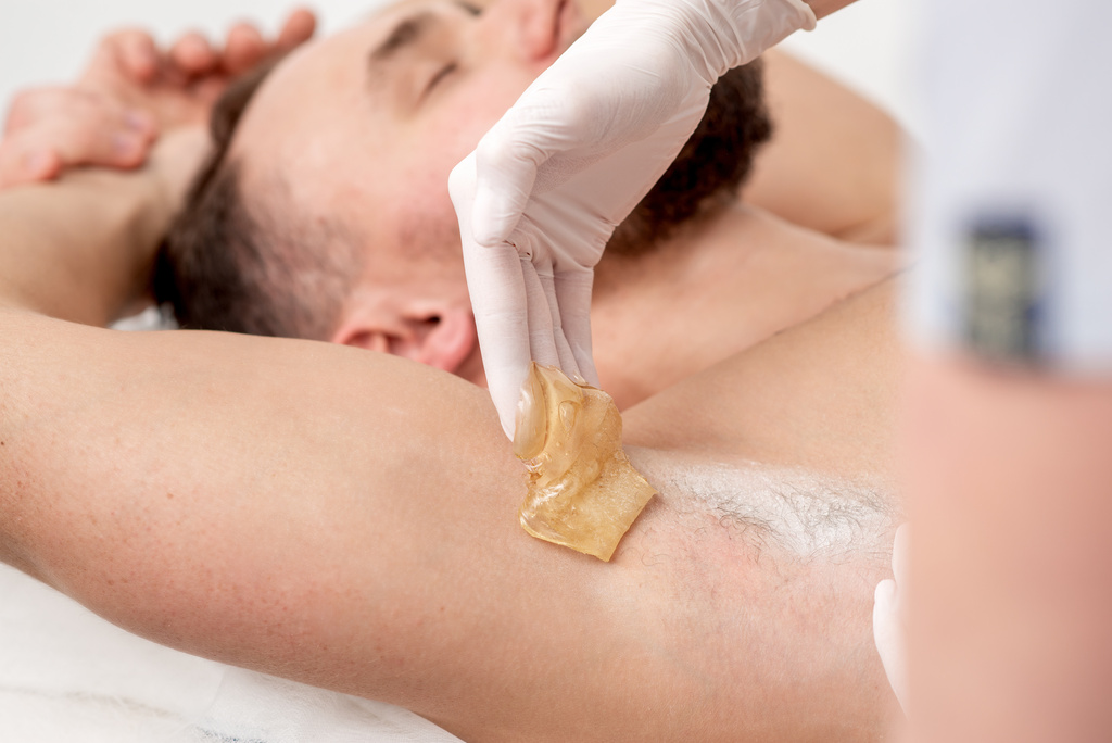 Mies makaa kauneushoitolan hoitopöydällä. Kosmetologi sokeroi hänen kainalokarvojaan.